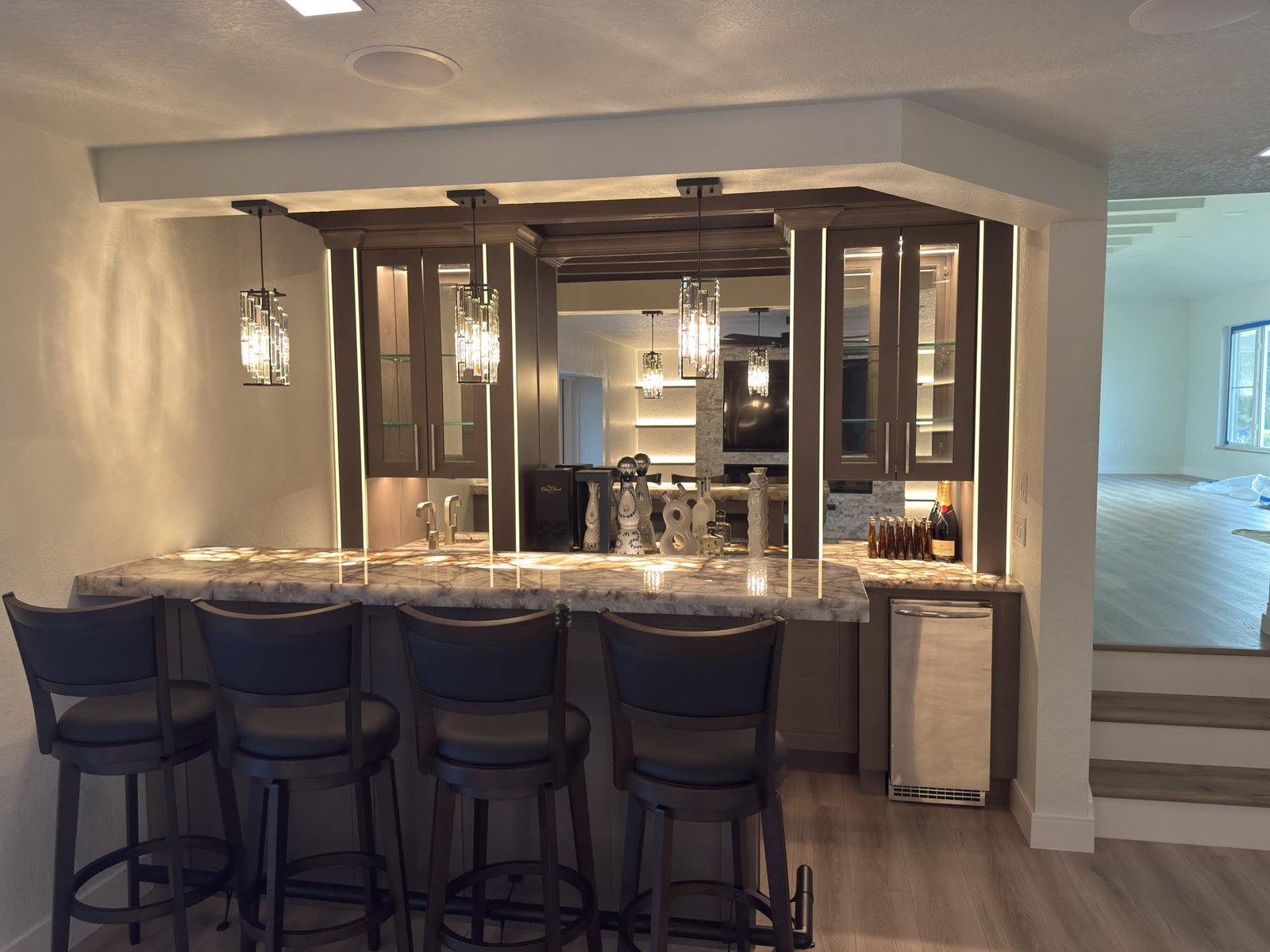 Custom home bar with a backlit stone counter, dark cabinets, glass-front display cases and crystal pendant lights