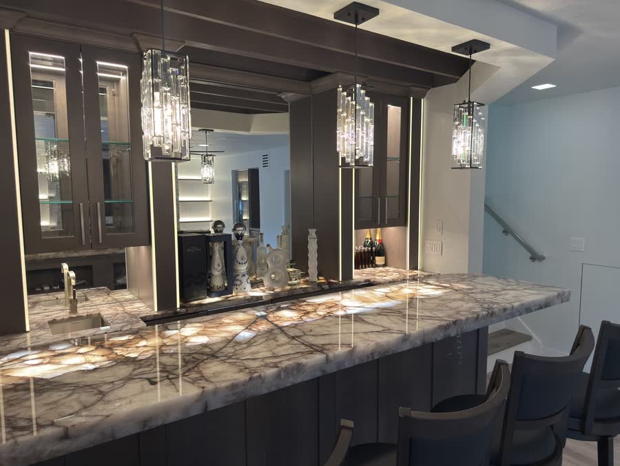 Home bar with a glowing backlit stone counter, lit glass display cabinets and crystal pendants