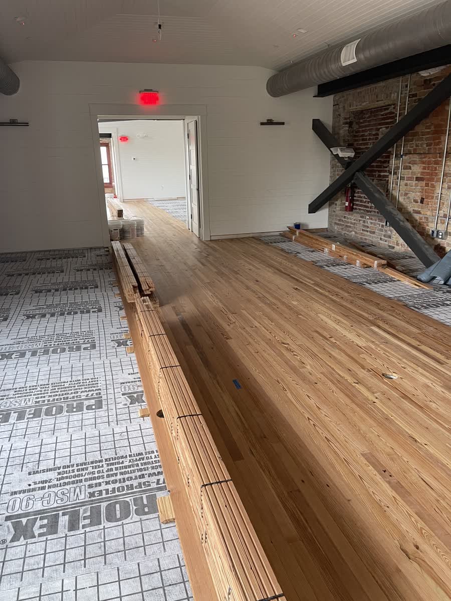 Solid wood floorboards being installed over underlayment along the brick wall